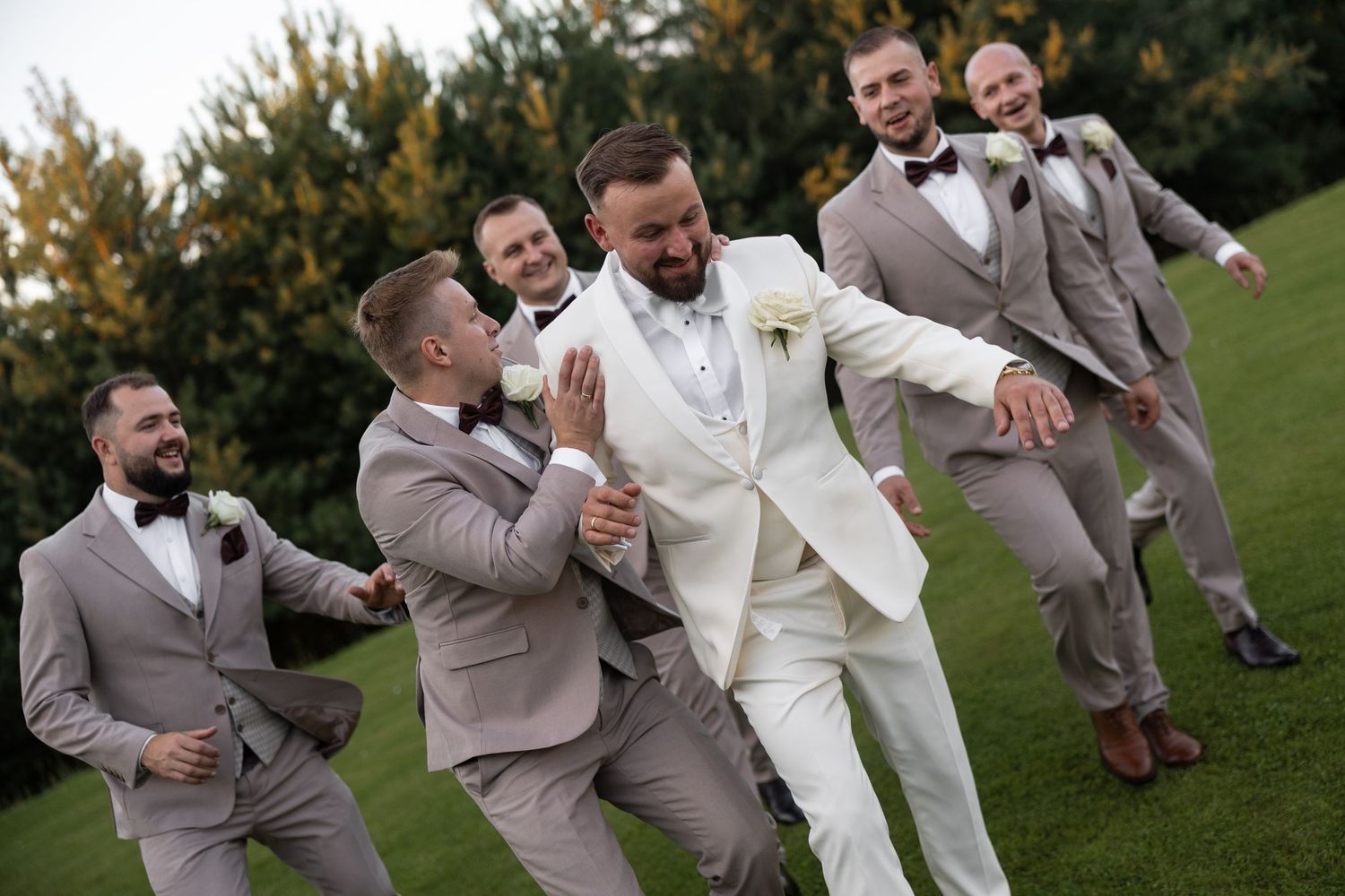 Groom in white suit laughing with groomsmen in tan suits and bow ties on green lawn at wedding.
