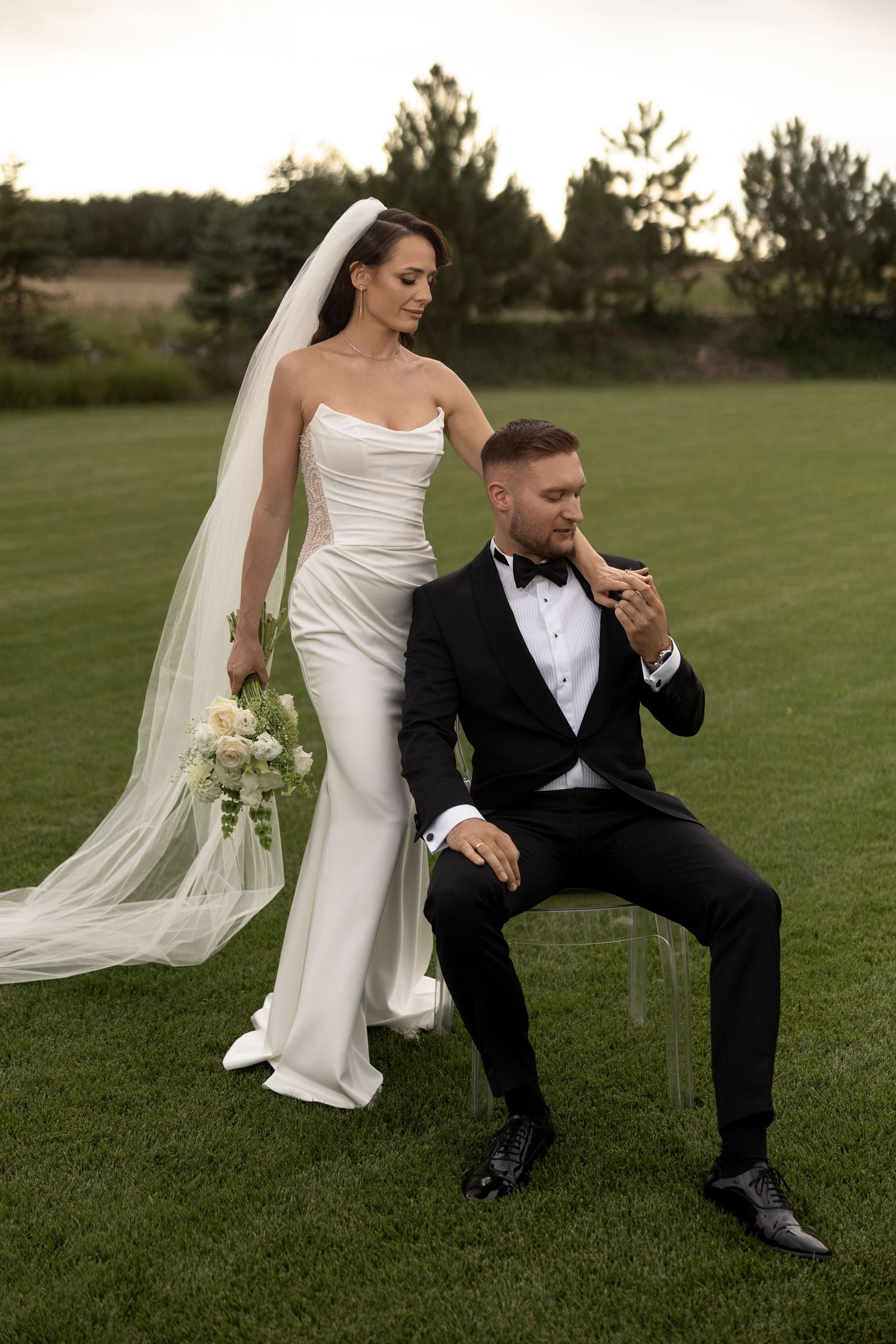 Elegant bride in white gown with veil holds bouquet beside groom in black tuxedo seated on chair outdoors.