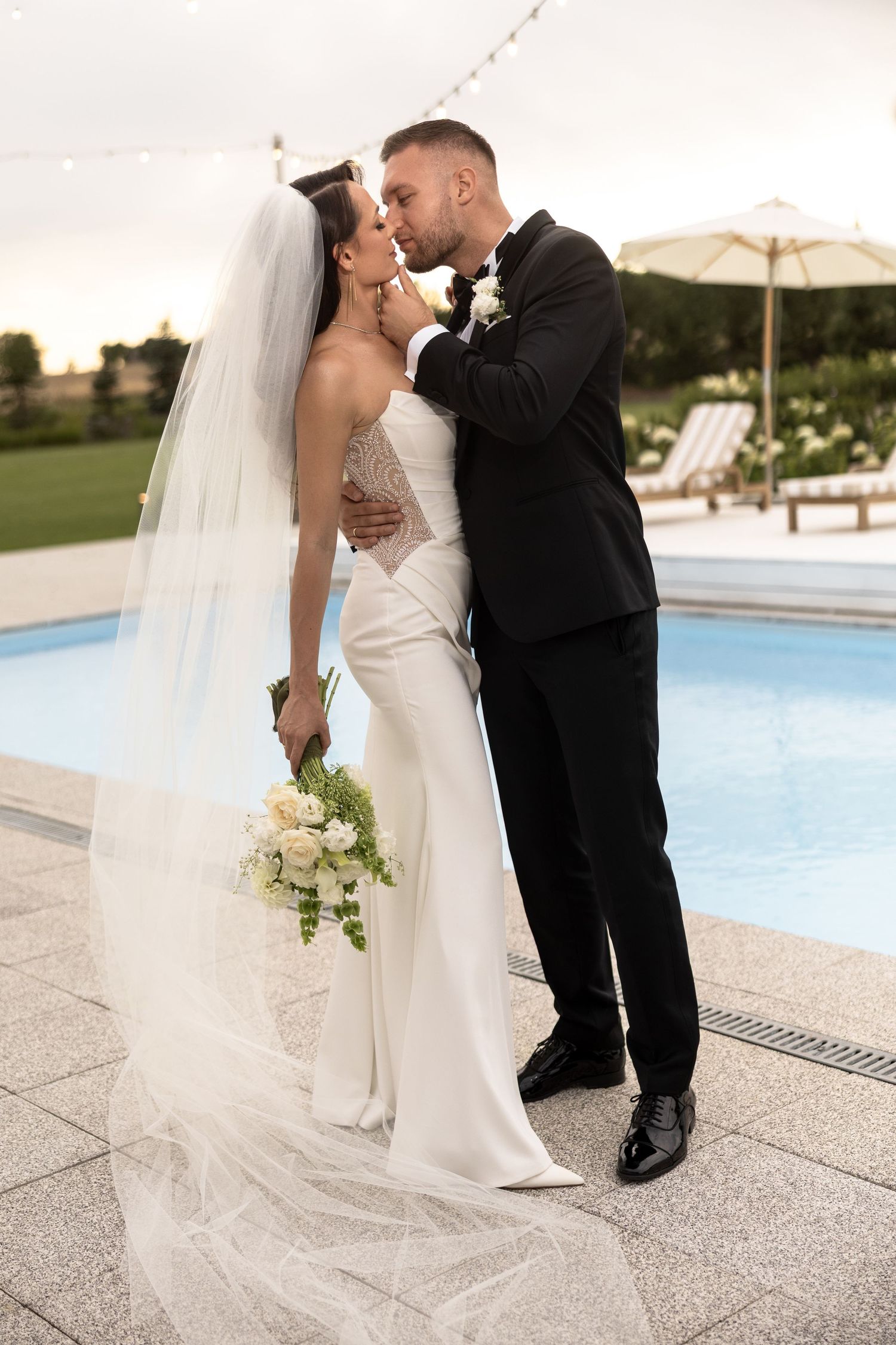 Bride and groom share a romantic kiss poolside, bride in white gown with veil holding bouquet, groom in black tuxedo.