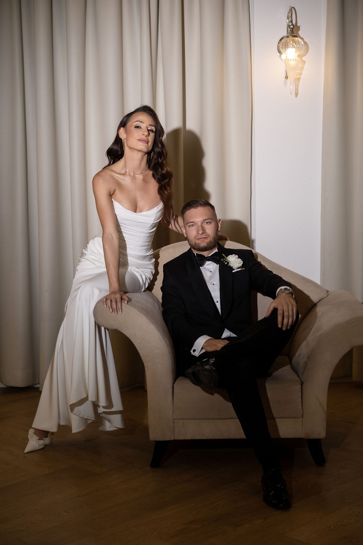 Bride in white gown and groom in black tuxedo pose elegantly in a softly lit room with cream curtains.