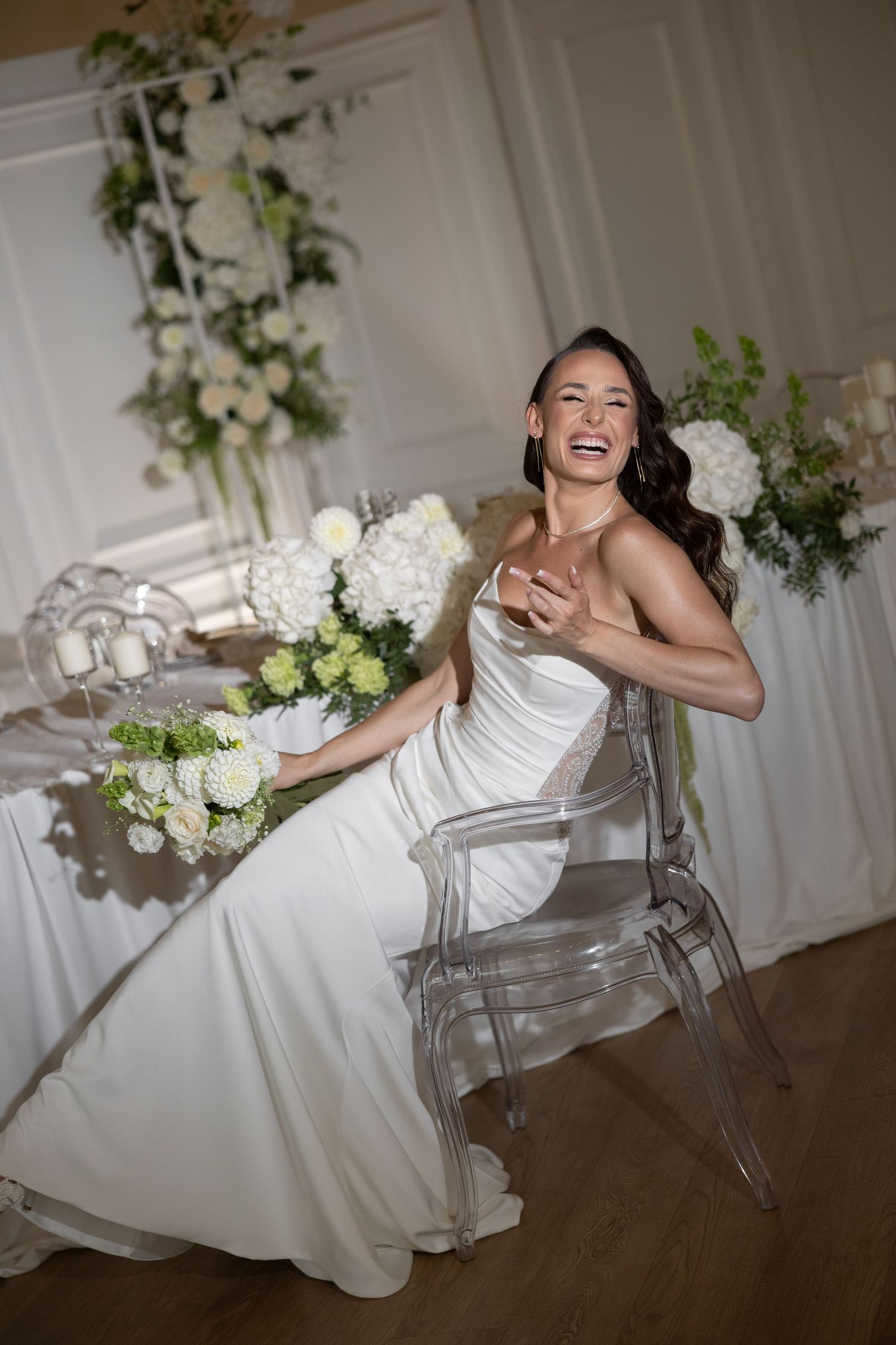 Laughing bride in white satin gown seated on acrylic chair surrounded by white hydrangea floral arrangements.