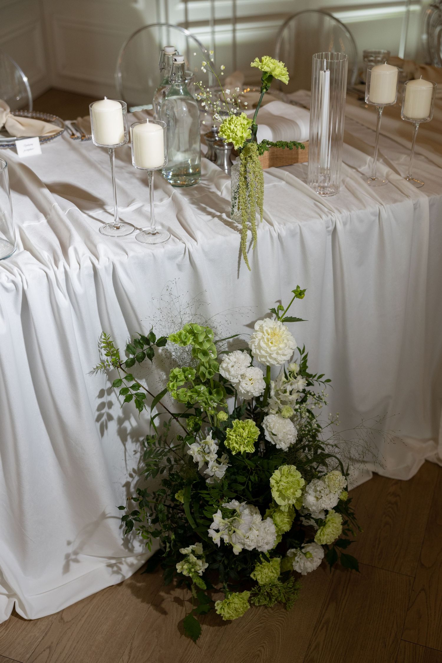 Elegant wedding table with white draping, candles, glass holders, and green-white floral arrangement on wooden floor.