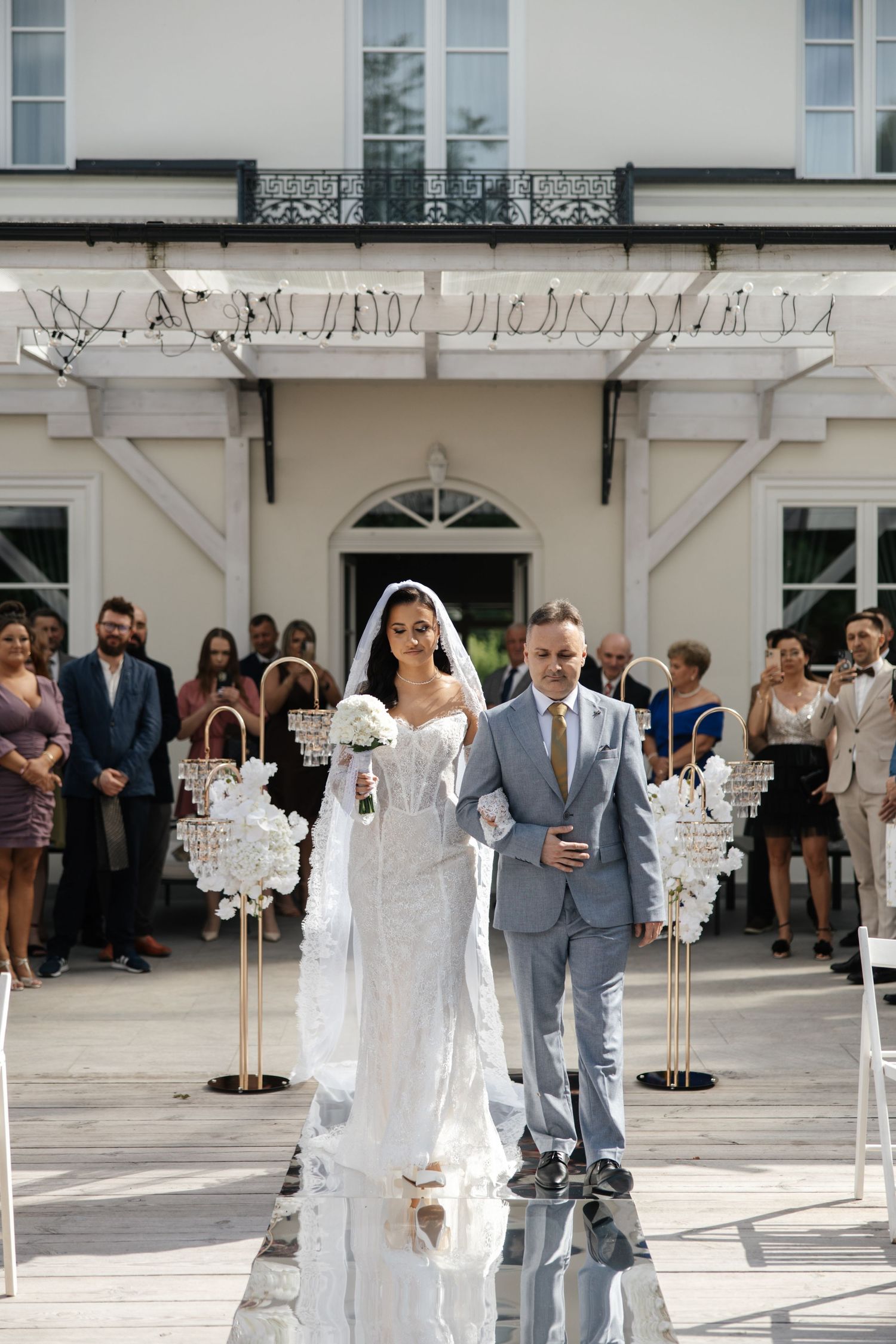 Bride in white gown walks down outdoor aisle with escort at elegant wedding ceremony, guests seated beside floral arrangements.