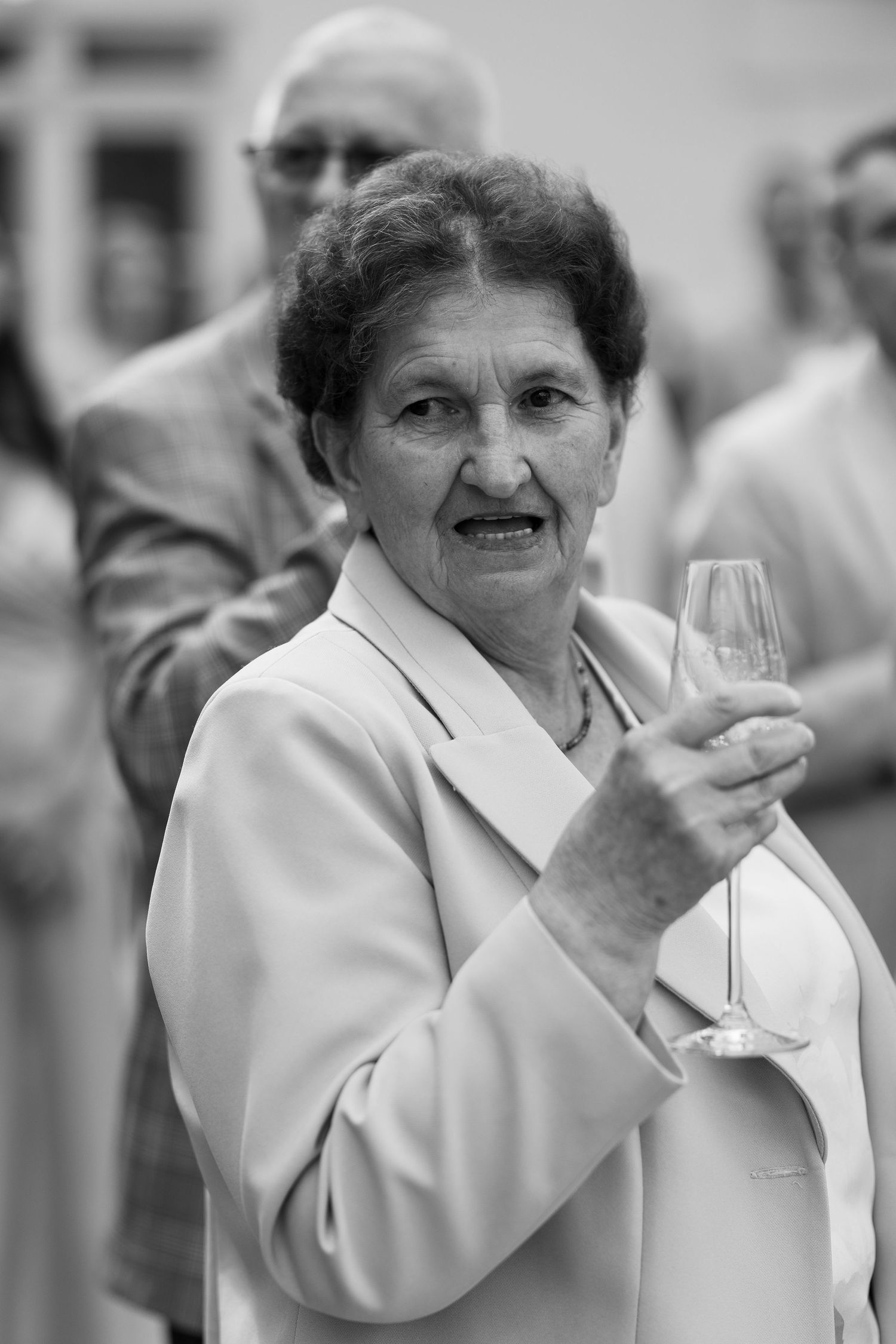 Elderly woman in light blazer holding champagne flute at formal event, black and white portrait photo.