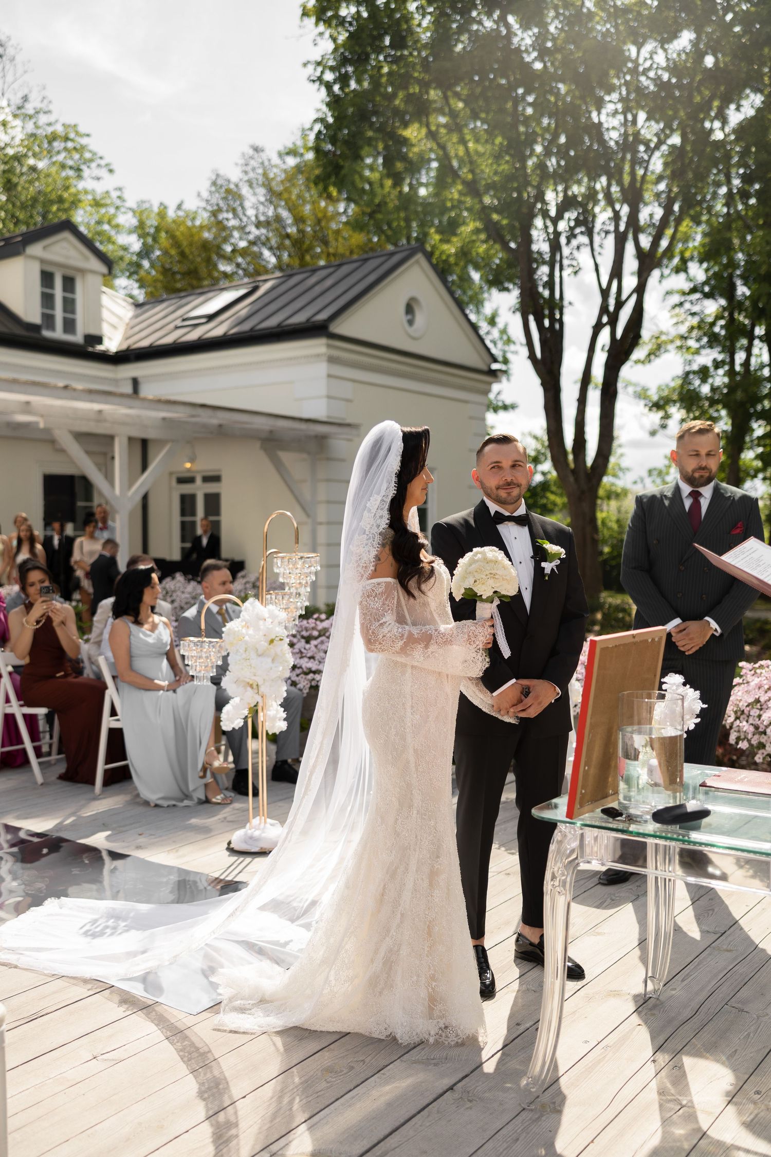 Bride and groom stand at outdoor wedding altar, bride in lace gown with veil holding white bouquet, officiant reads ceremony.