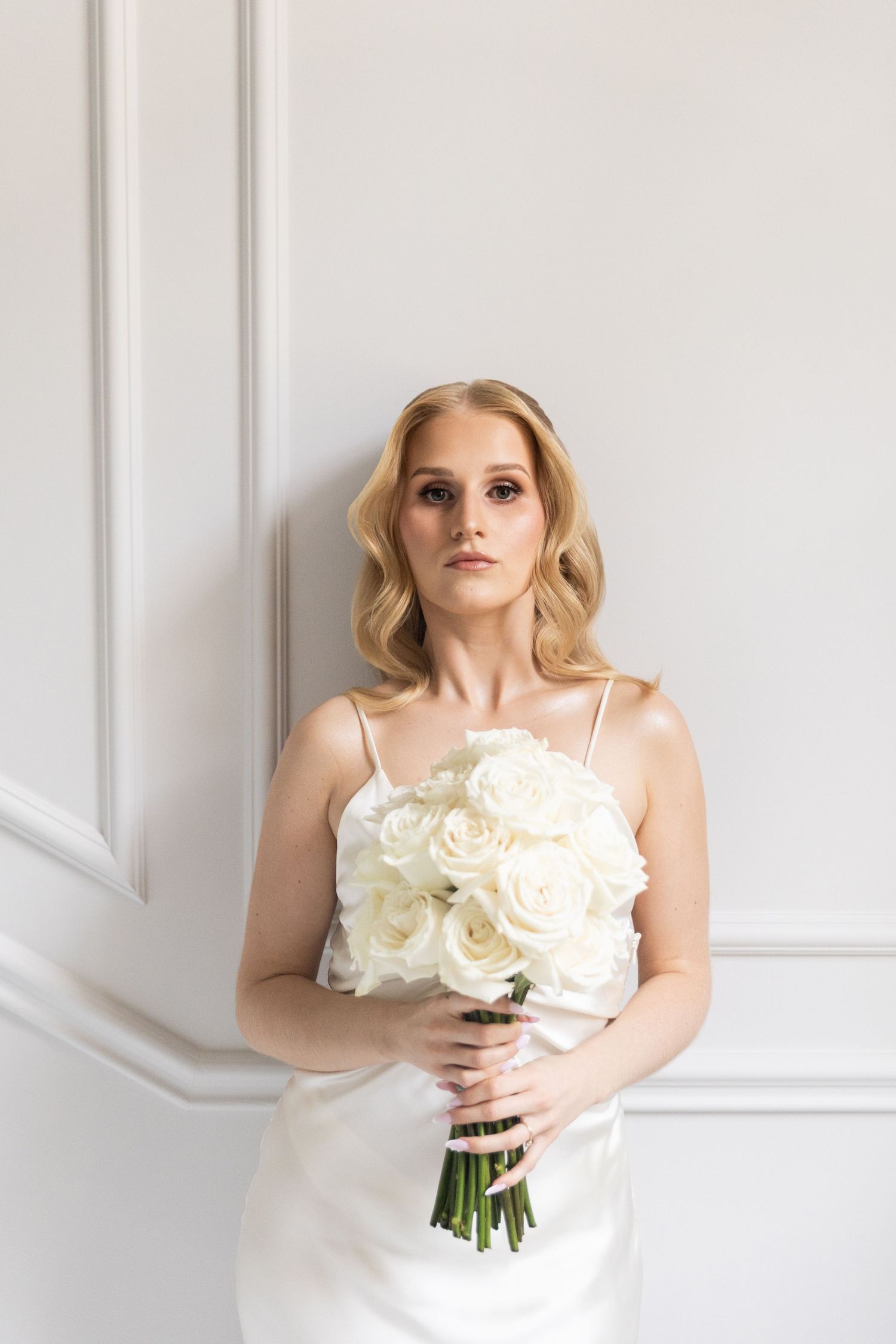 Elegant bride in white spaghetti strap dress holds classic bouquet of white roses against light wall.
