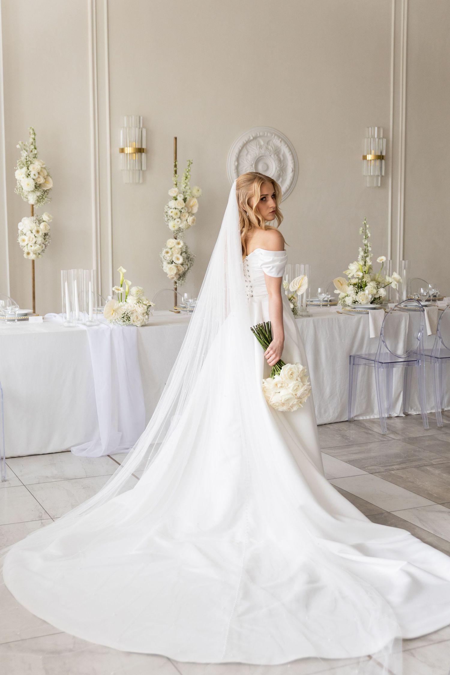 A flowing white wedding gown with a dramatic cathedral-length train displayed in a light-filled room with floral decor.