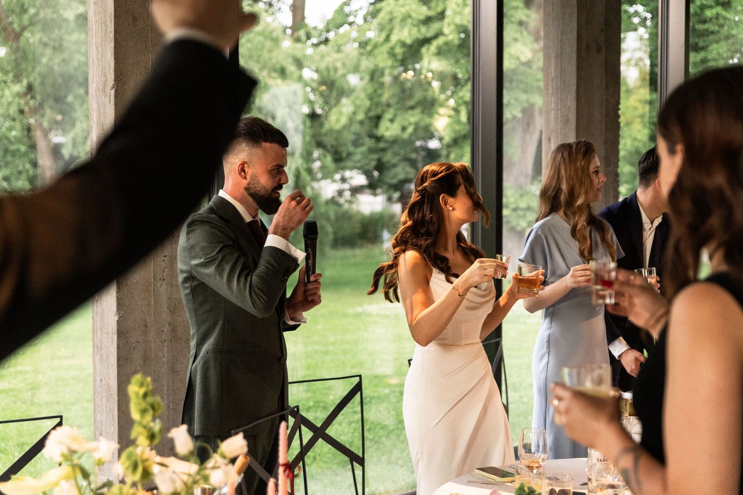 Wedding reception guests raise their glasses in a toast at a rustic outdoor venue under wooden beams and greenery.