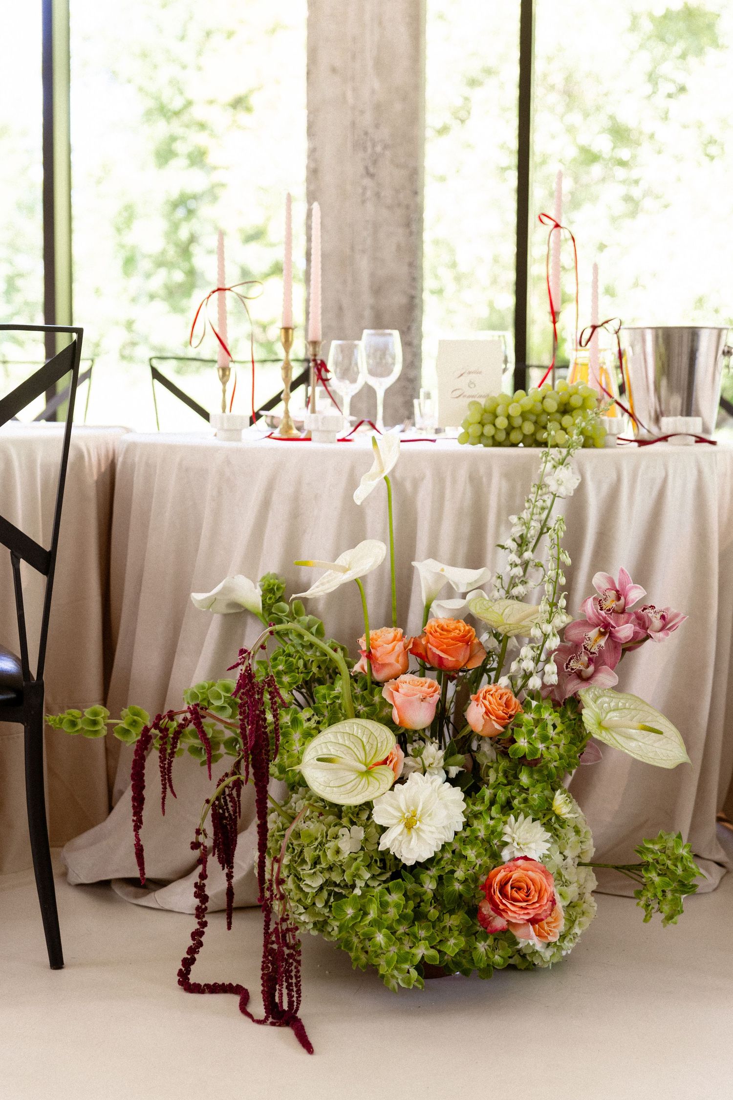 Elegant floral centerpiece with calla lilies, roses and grapes on a cream tablecloth at a formal dining setup.