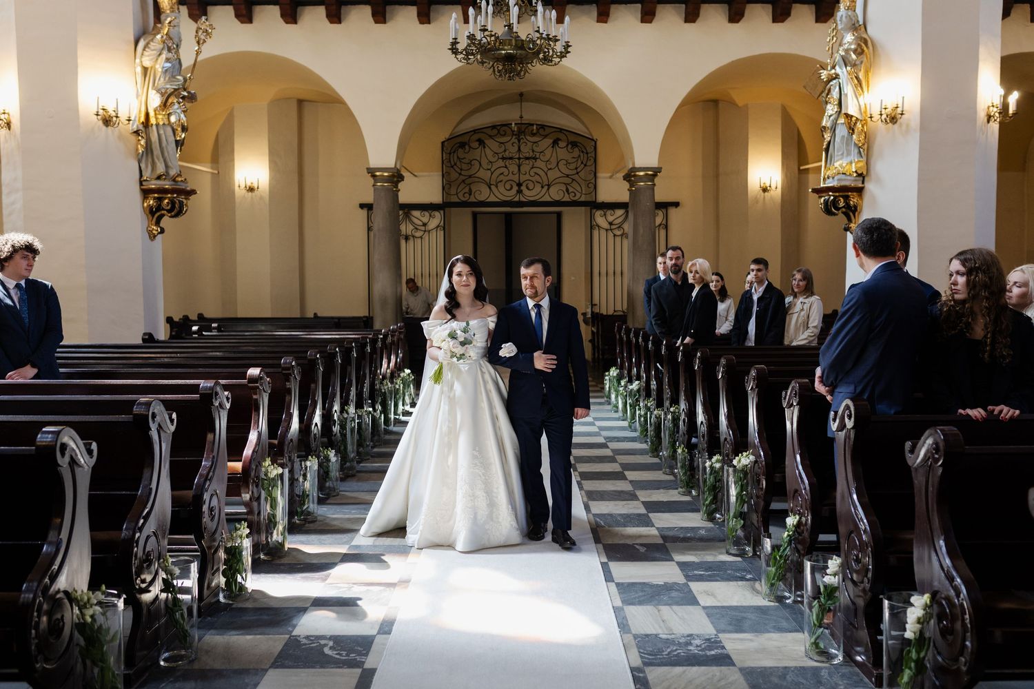 A wedding ceremony in a grand church with arched columns and a black and white checkered aisle runner.