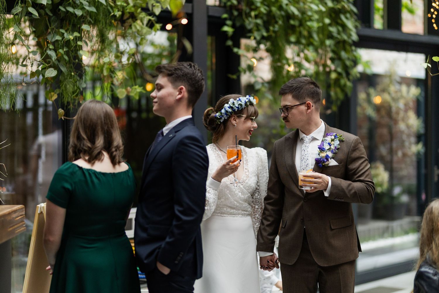 Wedding party members in formal attire gather outdoors near lush greenery and glass windows.