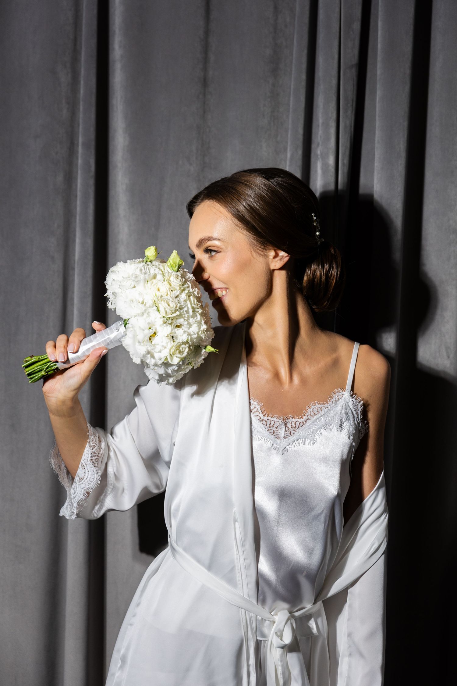 Bride in white silk robe and nightgown smells fresh white hydrangea bouquet in dramatic lighting.