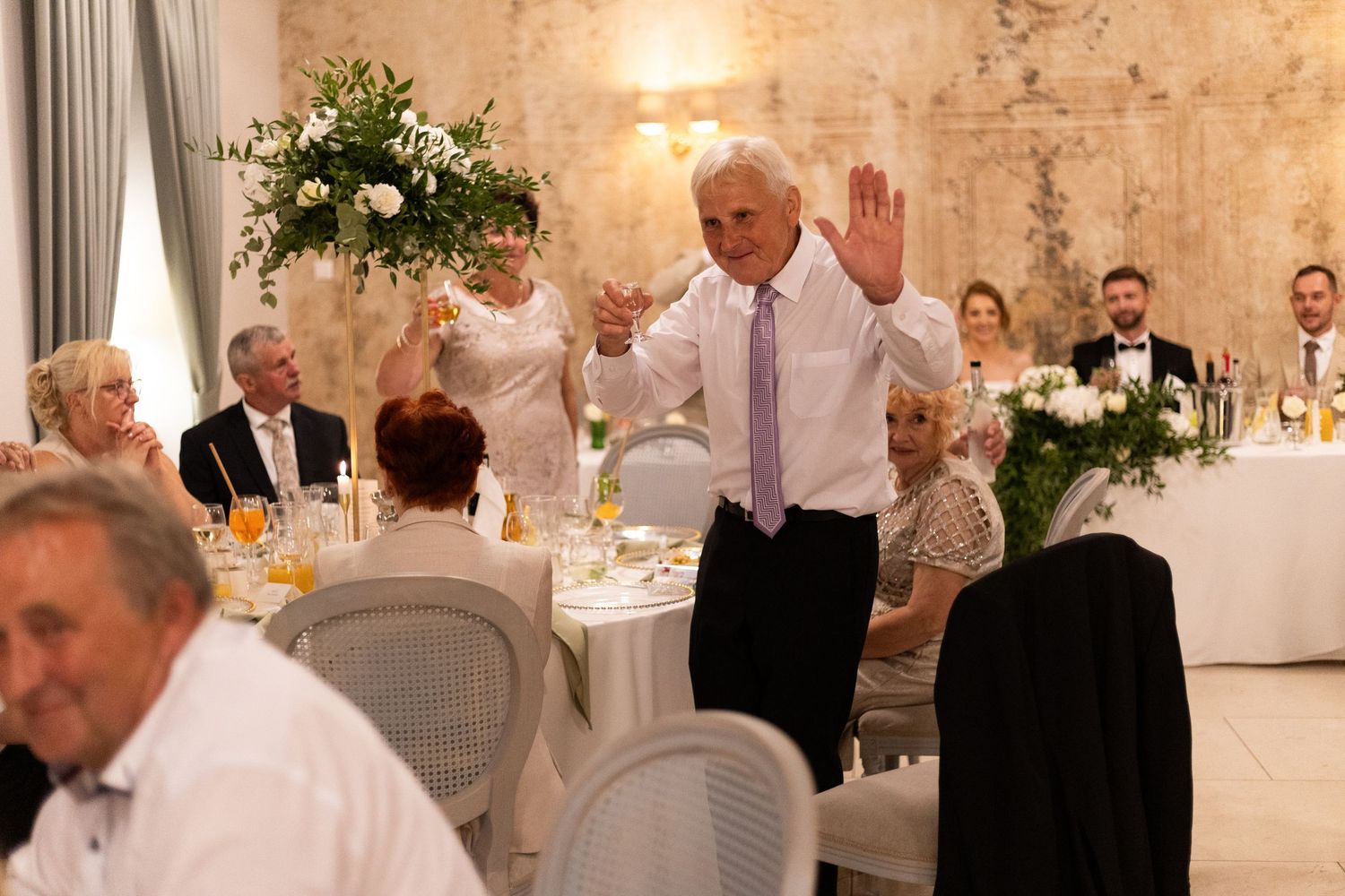 Wedding guests dance and socialize in an elegant reception hall with white decor and soft lighting.