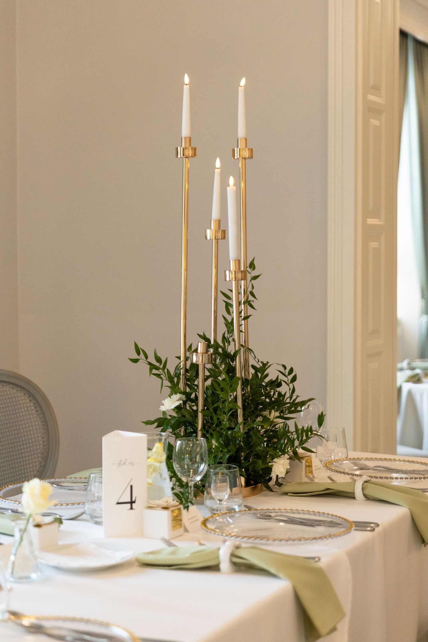 Elegant wedding table setting with tall gold candlesticks, white candles, greenery centerpiece and table number four.