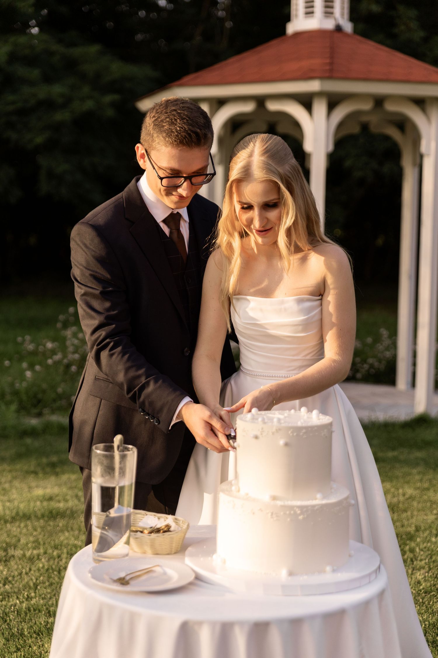 A couple in wedding attire cuts their white wedding cake together at an outdoor reception with a gazebo in background.