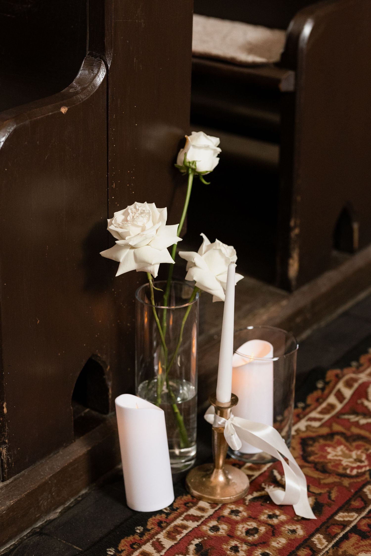 White candles and flowers in glass vases create an elegant ambiance along a dark wooden church pew.