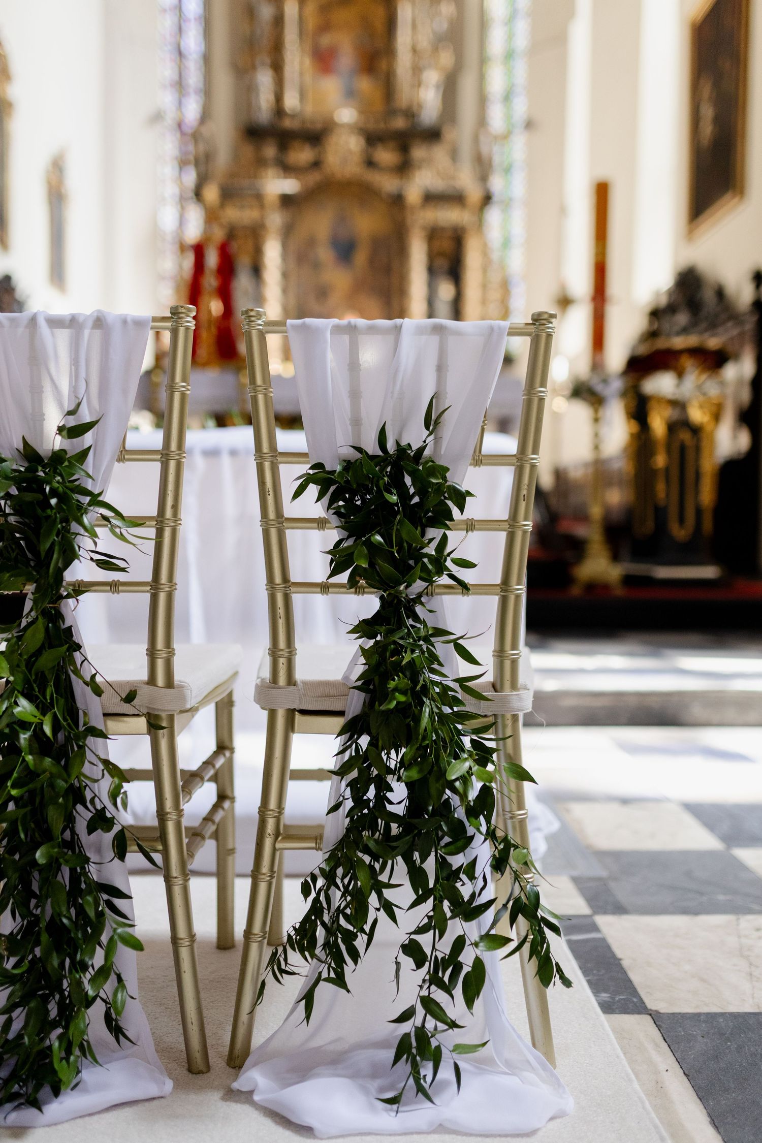 Elegant church ceremony chairs decorated with cascading greenery garlands and white fabric in a historic cathedral setting.