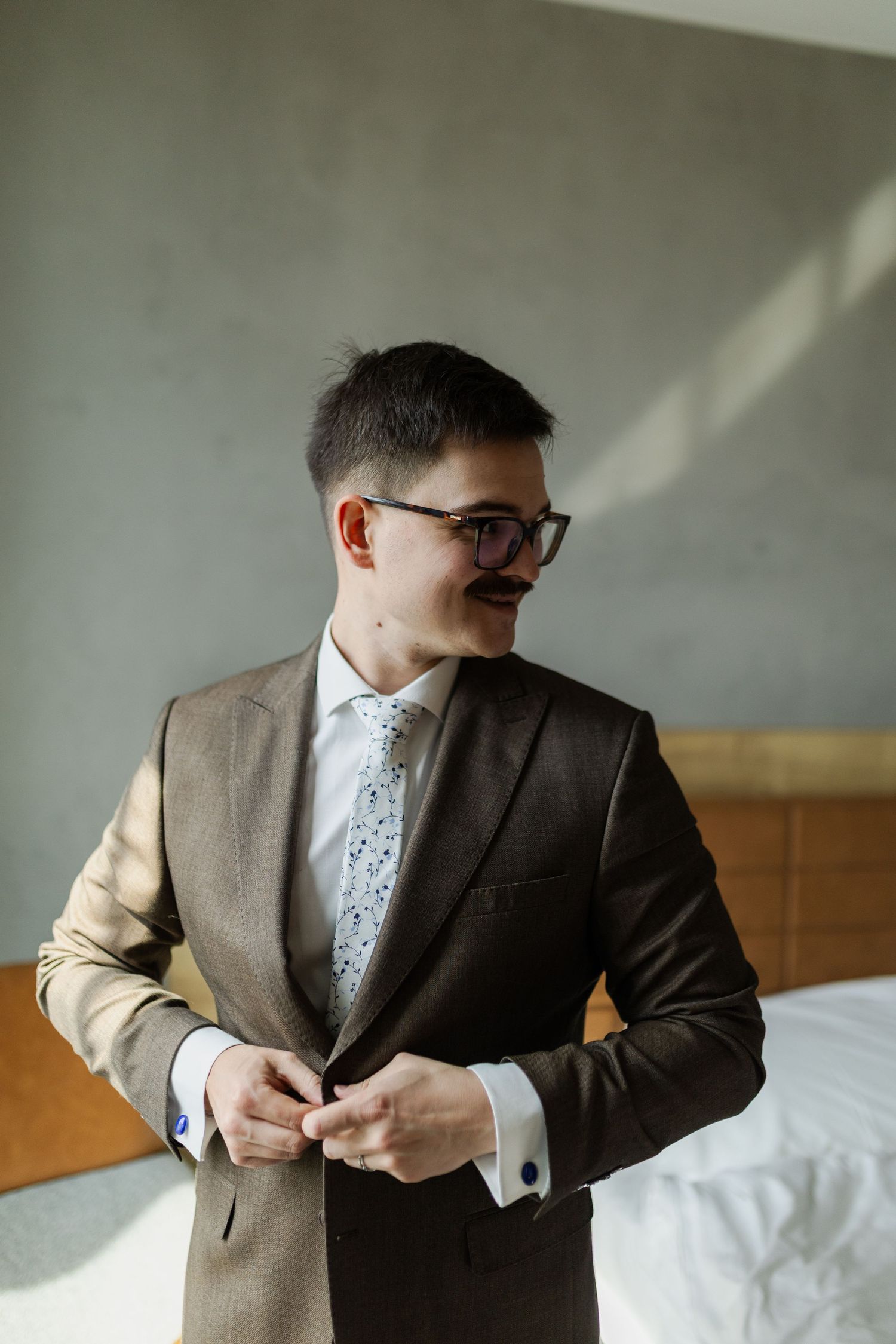 Groom in brown suit with glasses and floral tie buttons jacket in sunlit room with white walls and wooden furniture.