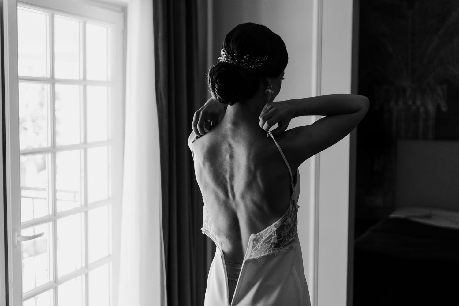 Black and white photo of elegant silhouette gazing through window in formal lace back dress.