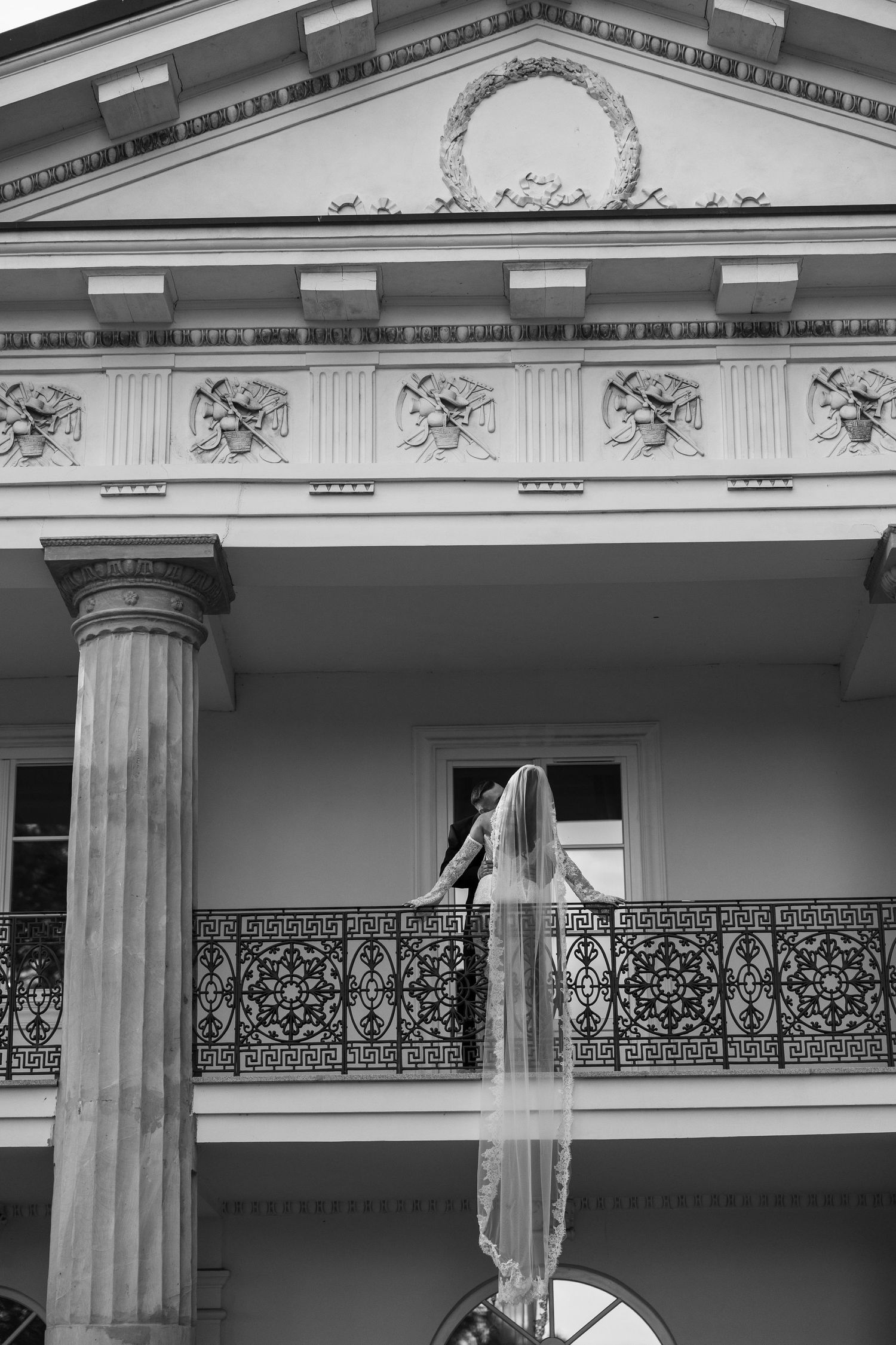 Black and white photo of a bride in a long veil standing on an ornate balcony of a classic Southern mansion.