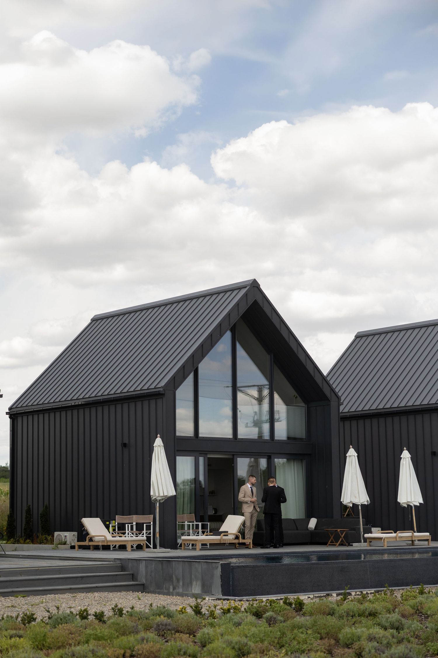 Modern black barn-style home with gabled metal roof, large windows, outdoor lounge area, and two people on the patio.