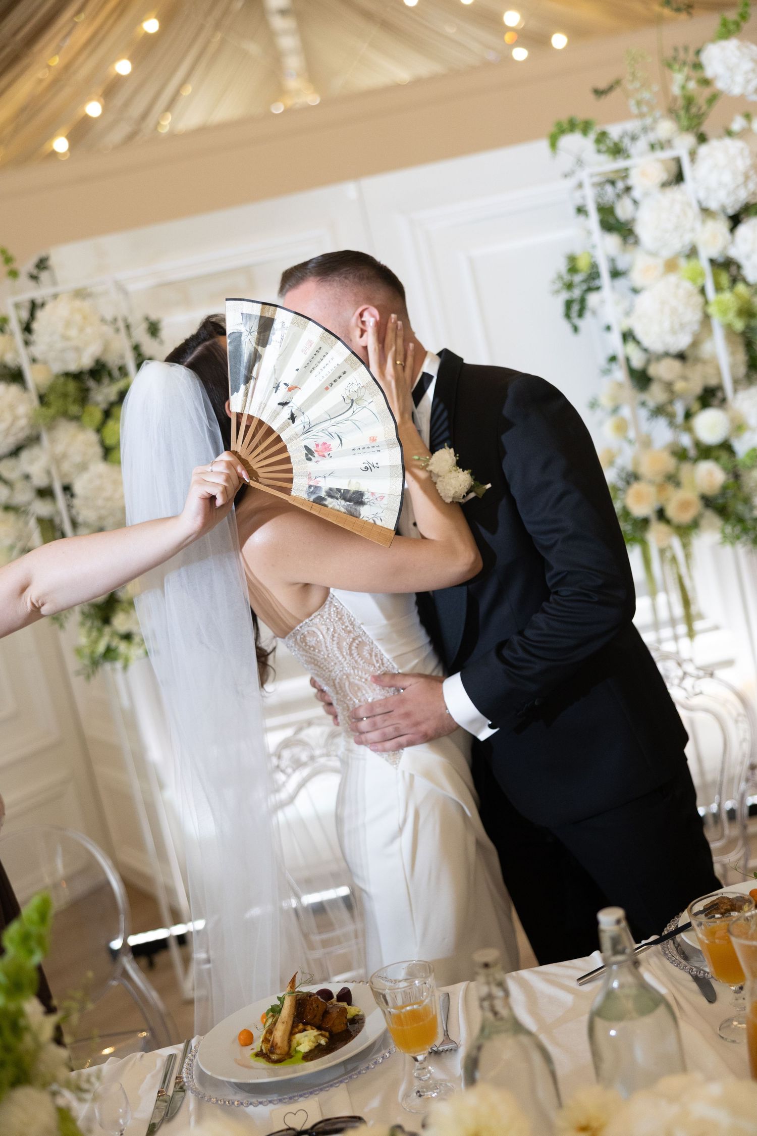 Bride and groom share a kiss hidden behind a decorative fan at their elegant wedding reception.