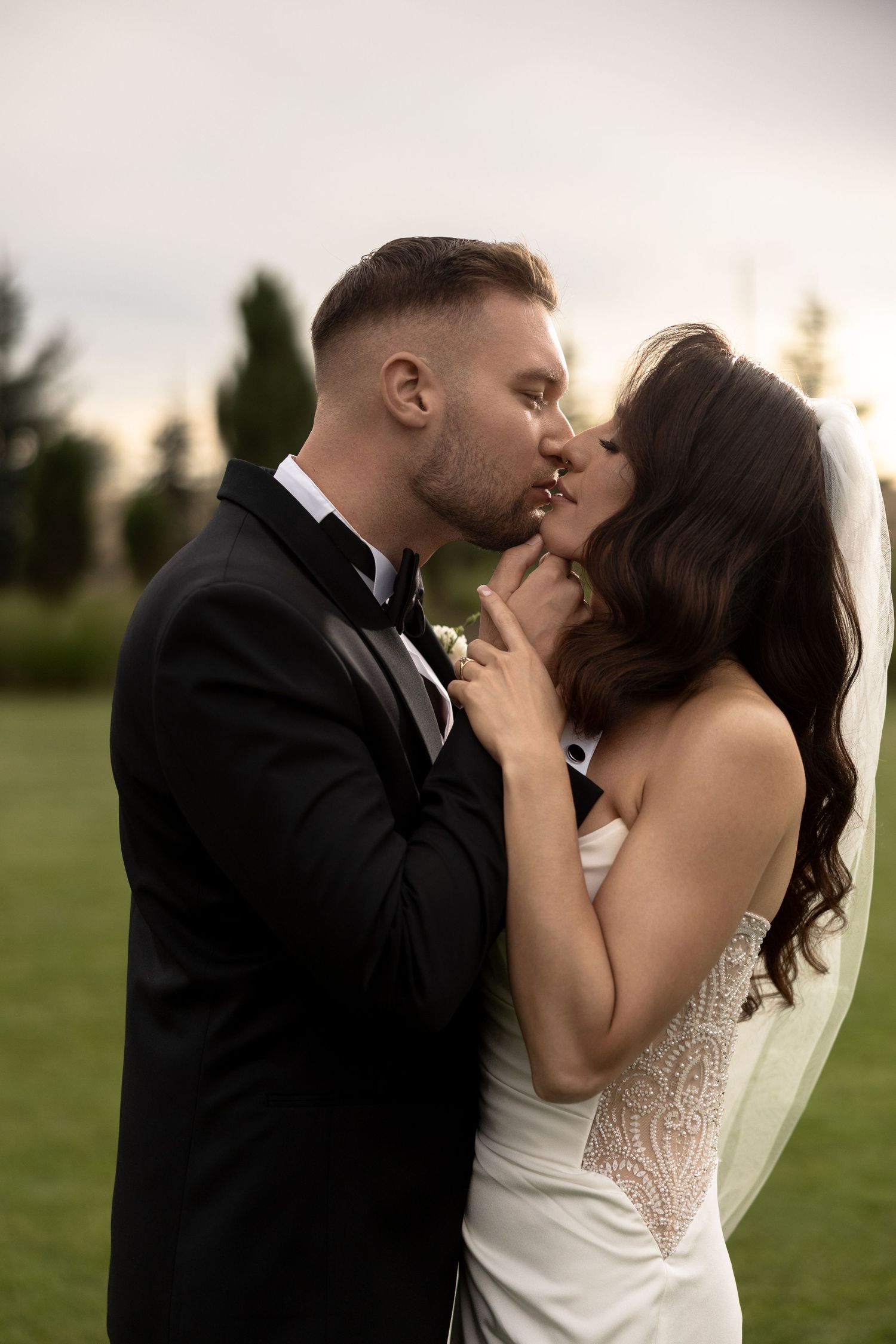 A bride and groom share a romantic kiss outdoors, dressed in wedding attire with a veil flowing behind them.