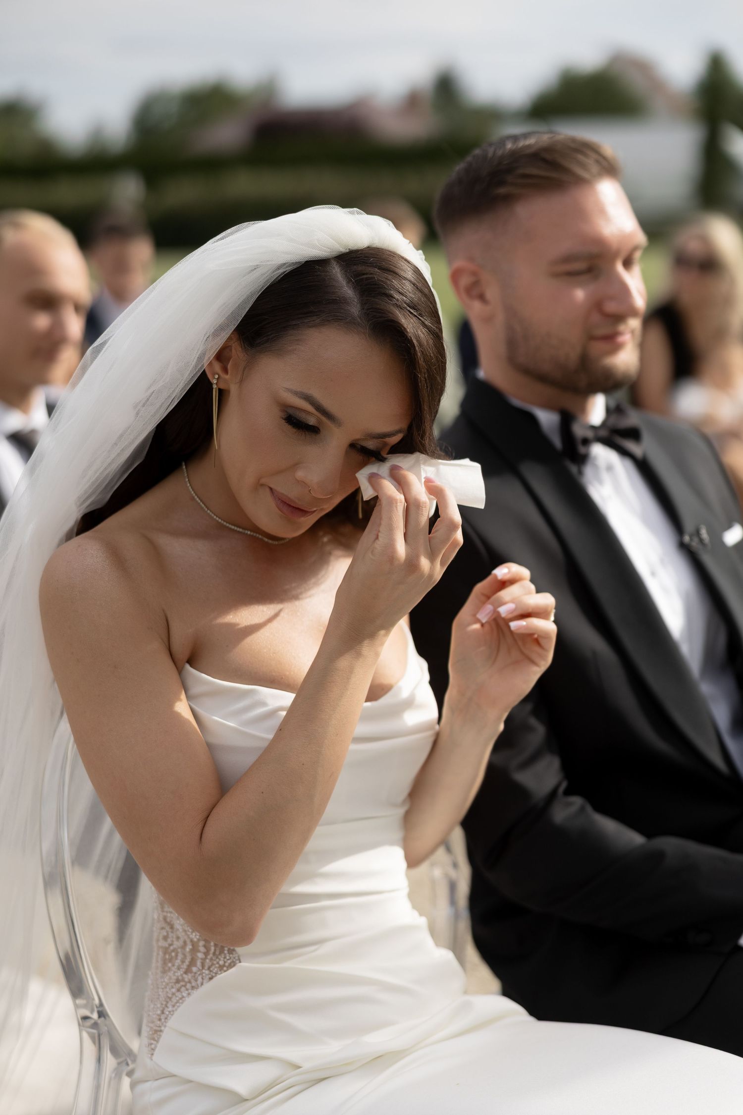 Emotional bride wiping tears with handkerchief during outdoor wedding ceremony, groom in tuxedo seated beside her.