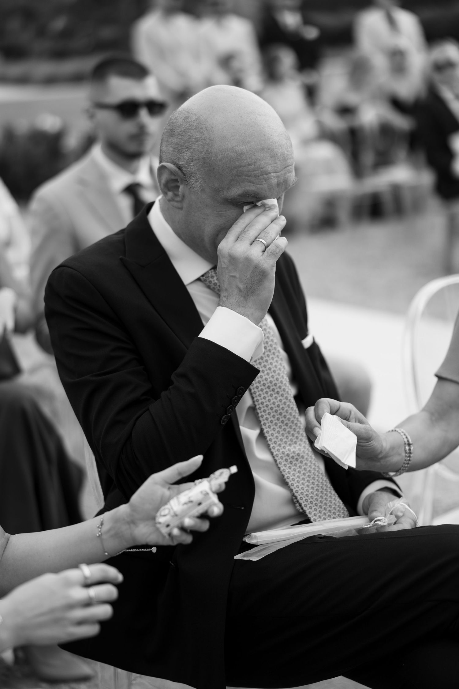 Emotional bald man in suit wiping tears at outdoor wedding ceremony, black and white photo.