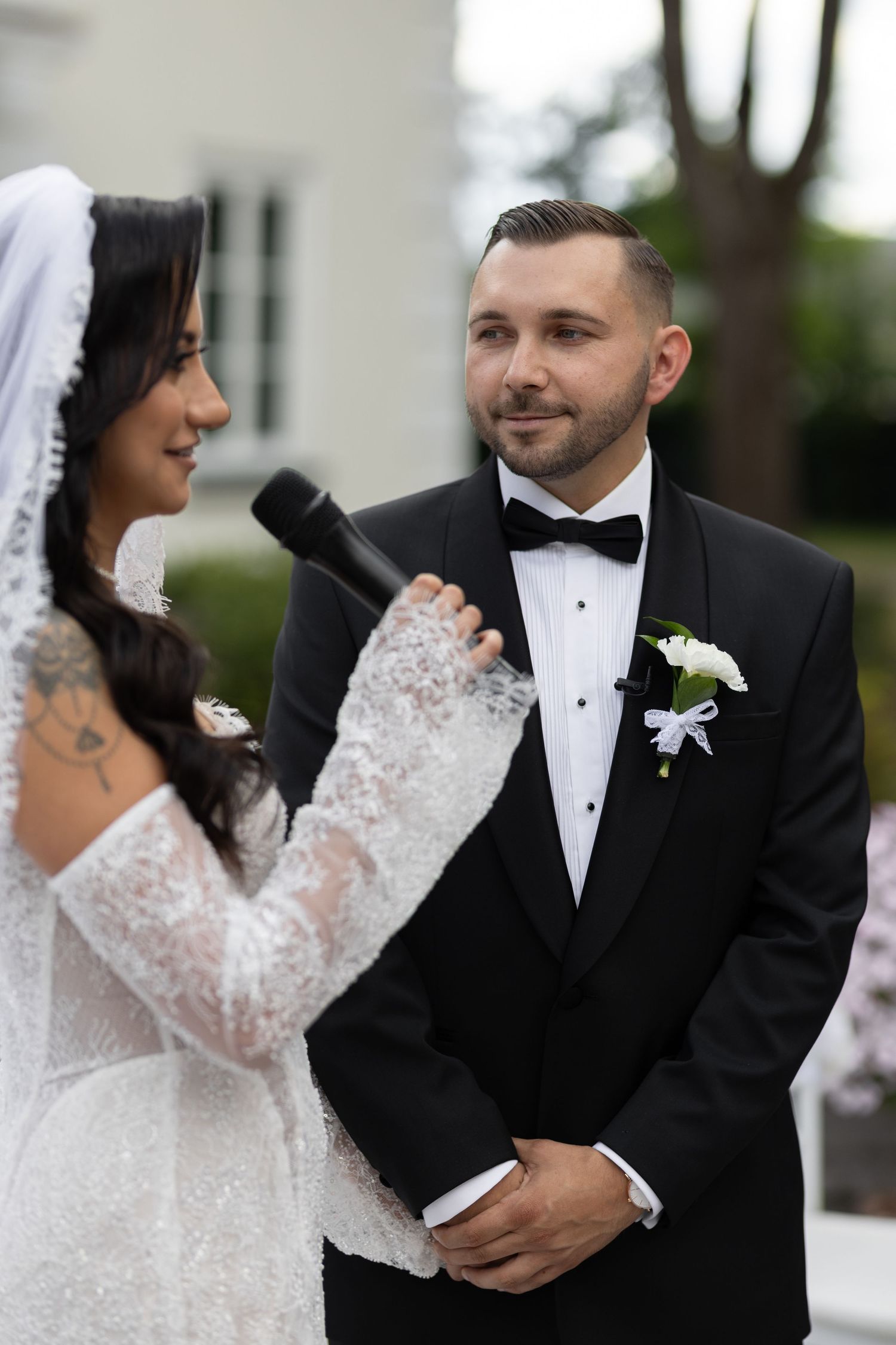 Bride in lace gown reads vows to groom in black tuxedo during outdoor wedding ceremony.