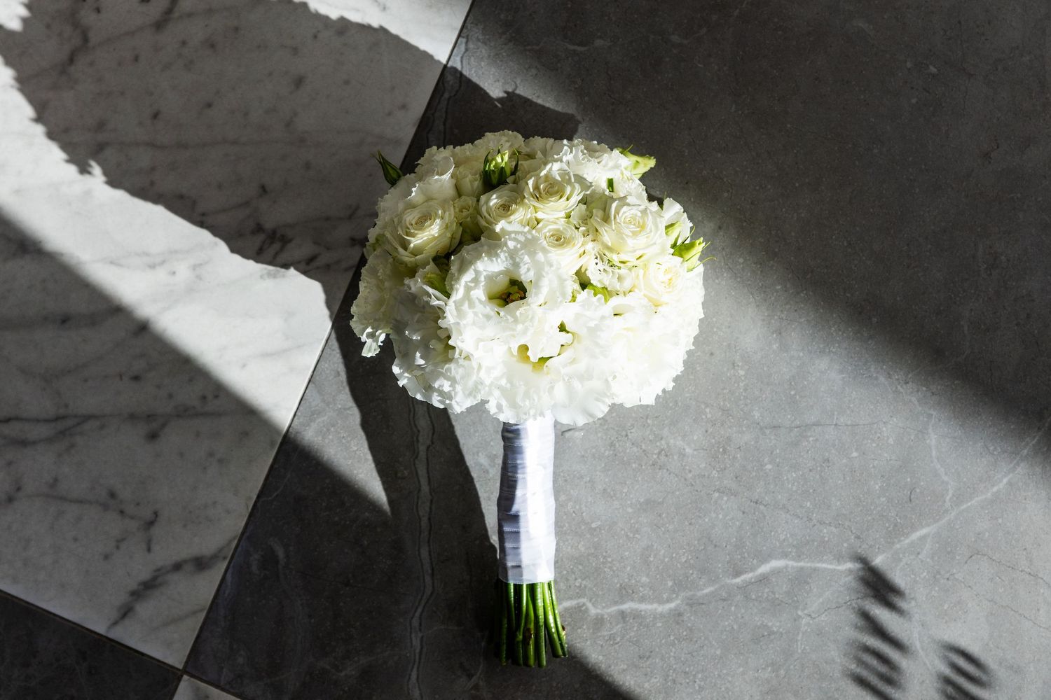 Elegant white floral bridal bouquet with green accents rests on marble surface in dramatic lighting.