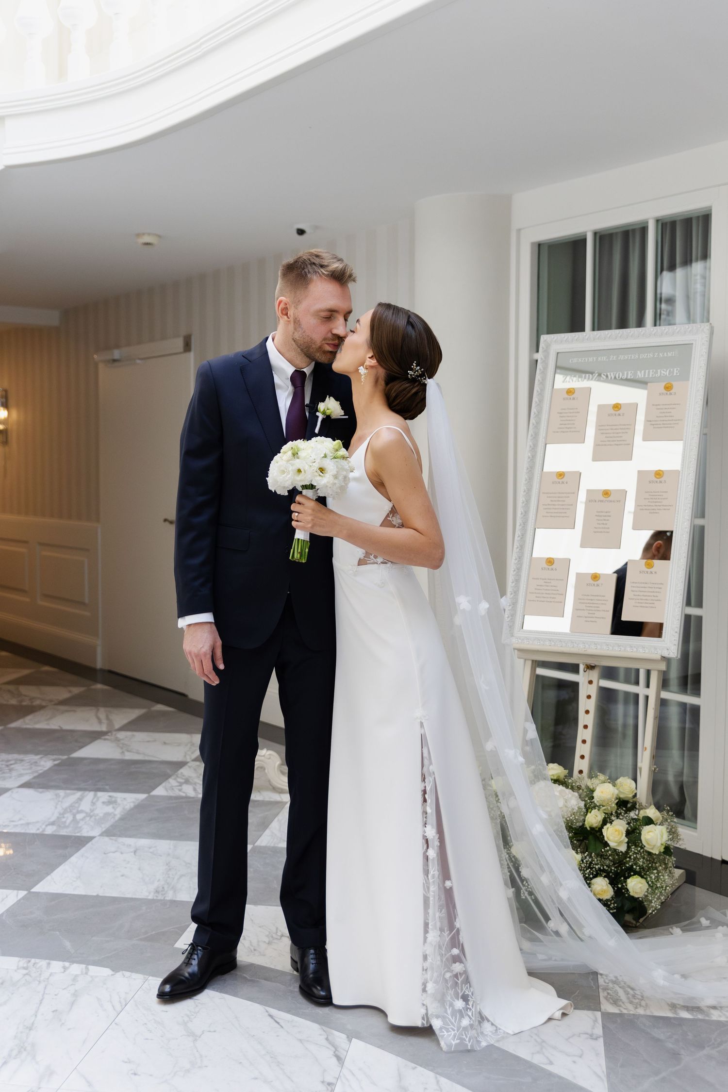 A bride and groom share an intimate moment in a hallway, with a wedding seating chart and floral decorations visible.