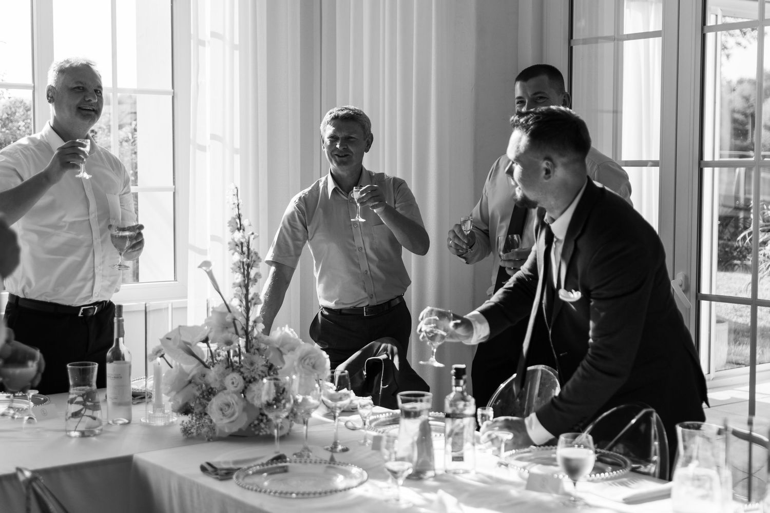 Black and white photo of people enjoying drinks and conversation at an elegant table setting with floral centerpiece.