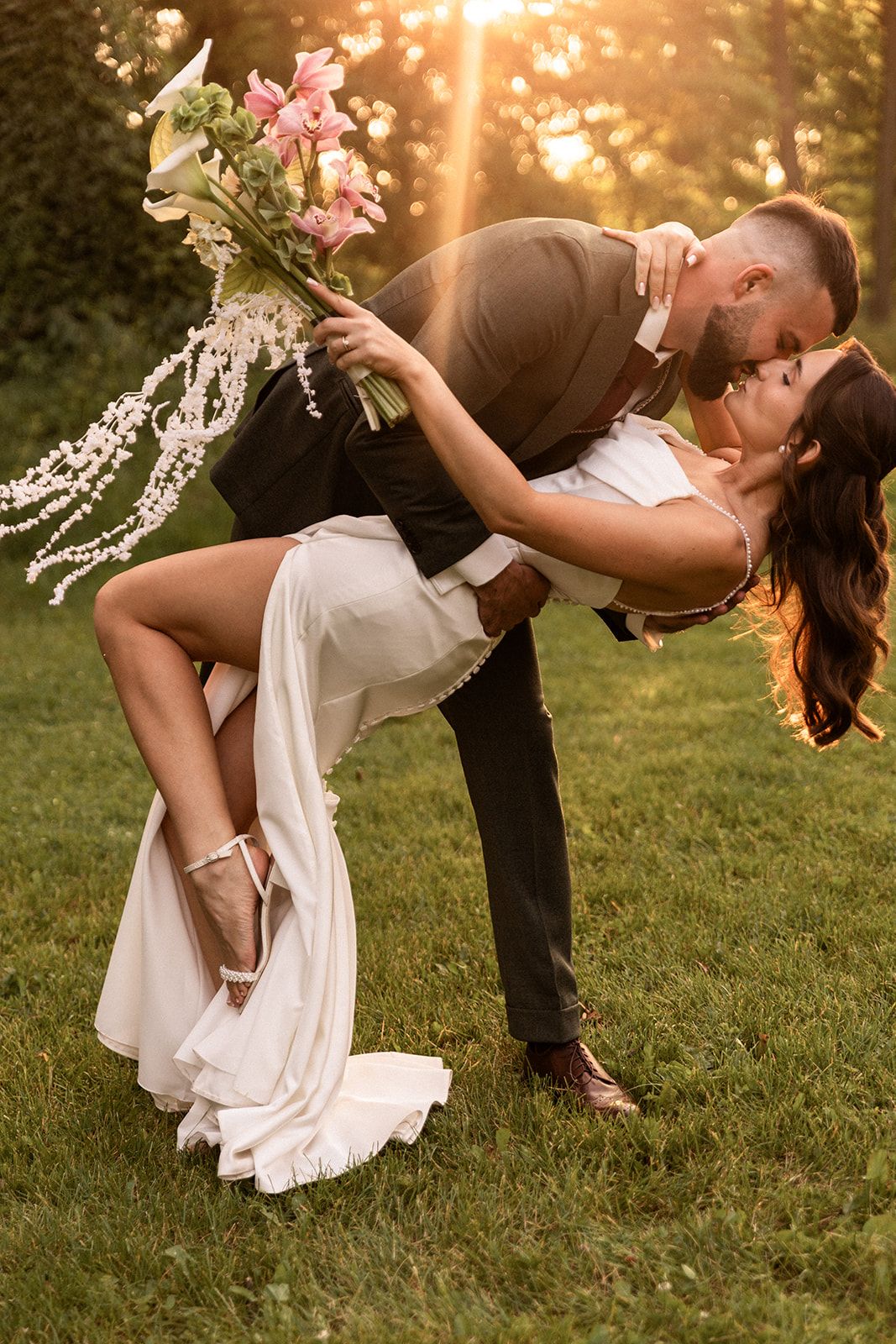 A romantic couple in wedding attire shares a passionate dip kiss at sunset in a grassy outdoor setting.