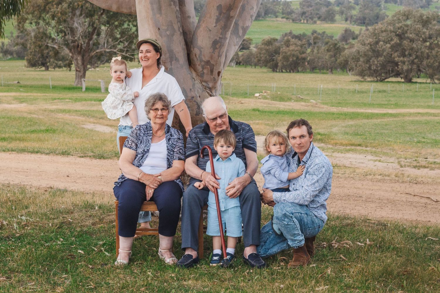 Family group sitting together outdoors under large eucalyptus tree in rural setting.