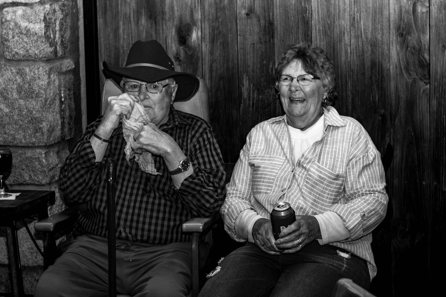 Two companions in cowboy hats share drinks and laughs while sitting against a wooden wall.