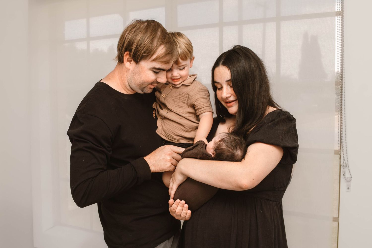 A loving family moment with parents in black clothing embracing their small child against a white wall.