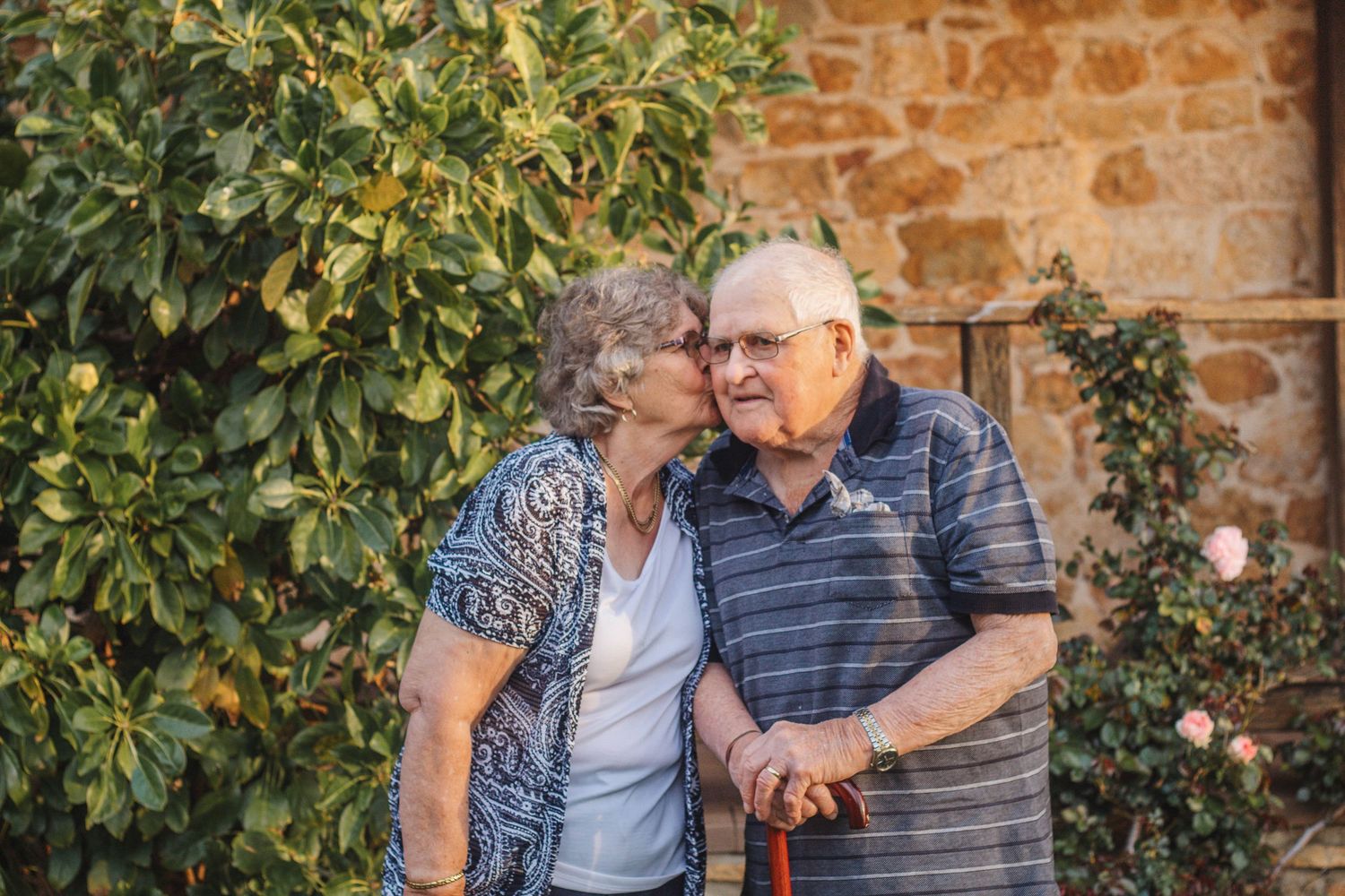An elderly couple shares a tender moment in front of a rustic stone wall with green foliage.