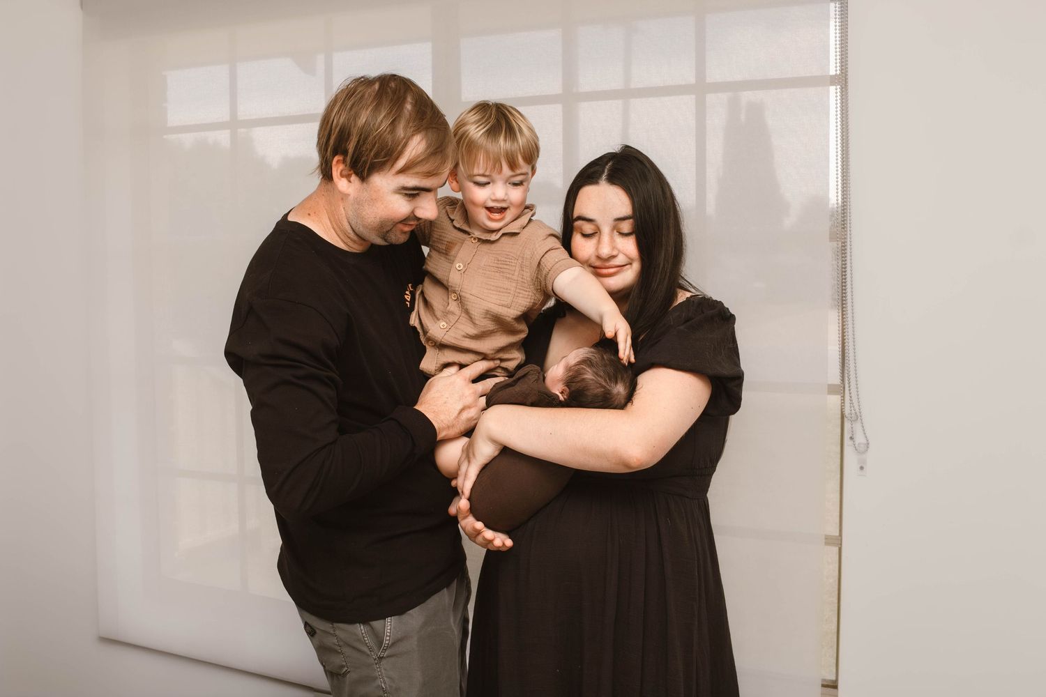 Parents in dark clothing share joyful moments with their toddler against a minimal white background.
