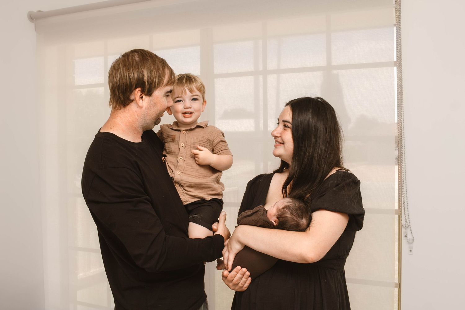 Warm family portrait showing parents in dark clothes sharing laughs with their child in brown clothing.