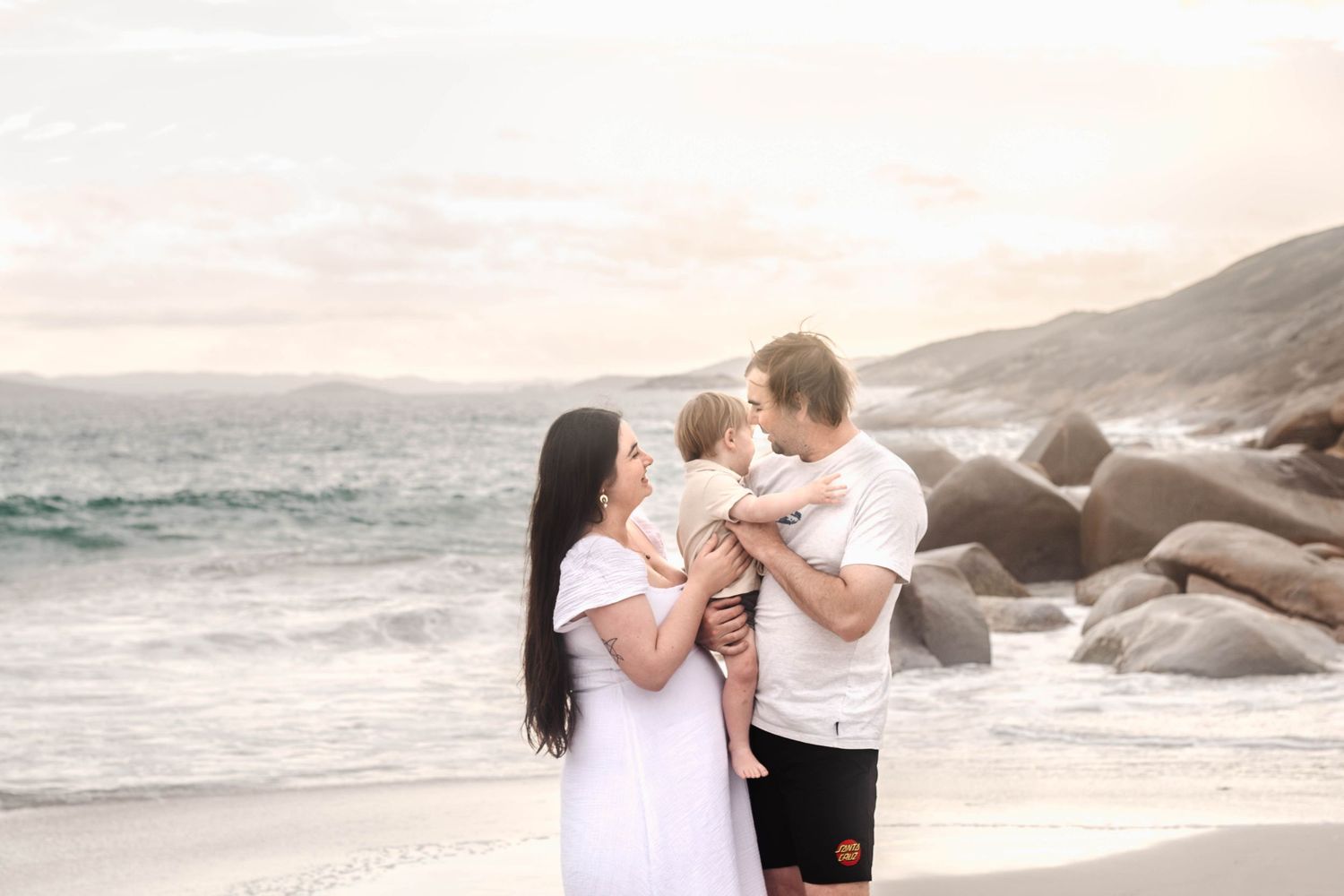 A couple shares a romantic dance pose on a scenic beach with rocky coastline at sunset.