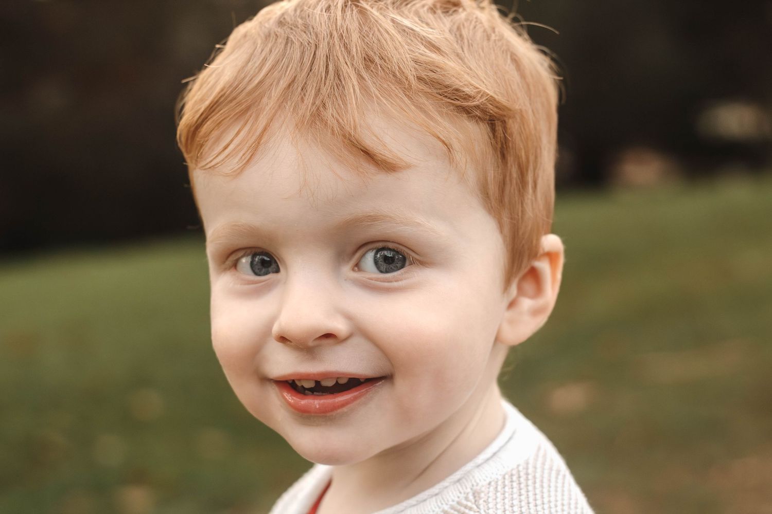 Young child with red hair smiles while standing outdoors wearing a white sweater.