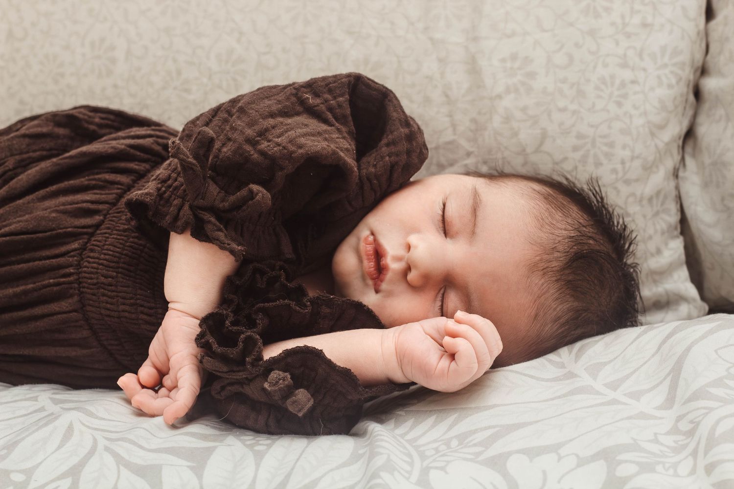 Peaceful sleeping baby wearing brown quilted dress on light bedding.