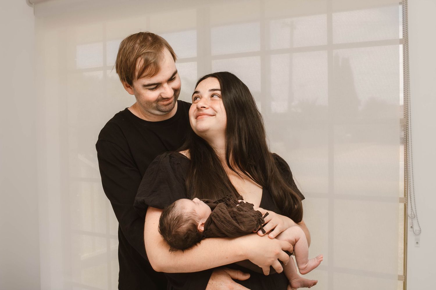 A loving couple embraces while holding their newborn baby in a bright, minimalist setting.