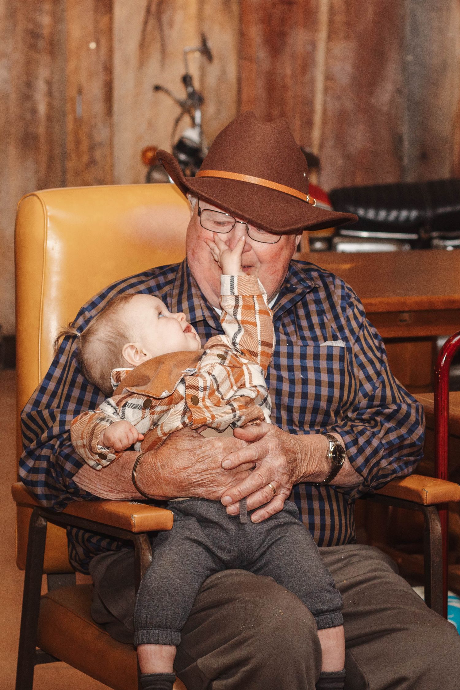 Person in cowboy hat and plaid shirt holding small child in leather chair.