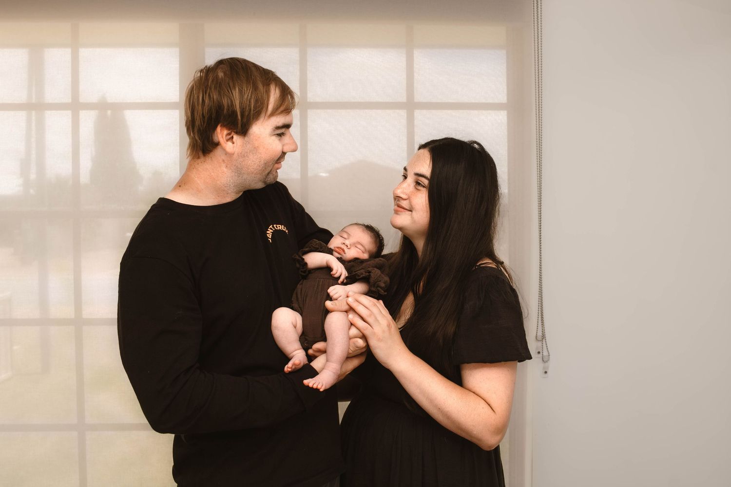 New parents share tender moments with their newborn baby against a bright window backdrop.