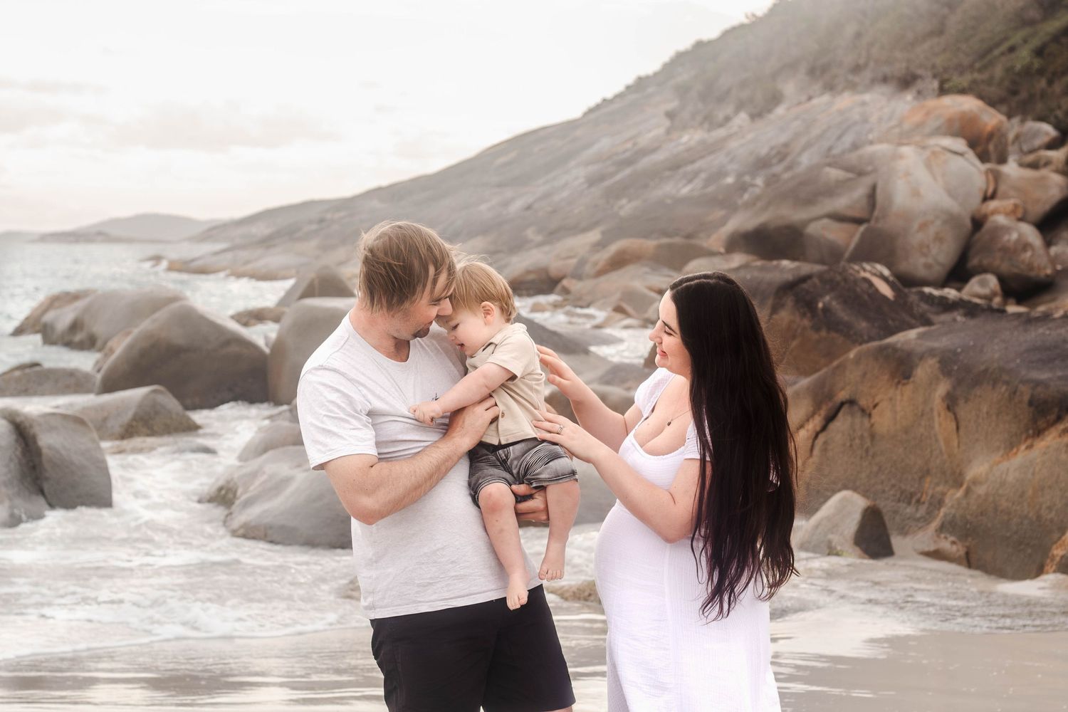 Parents playing with their baby on a scenic beach with rocky cliffs in the background.