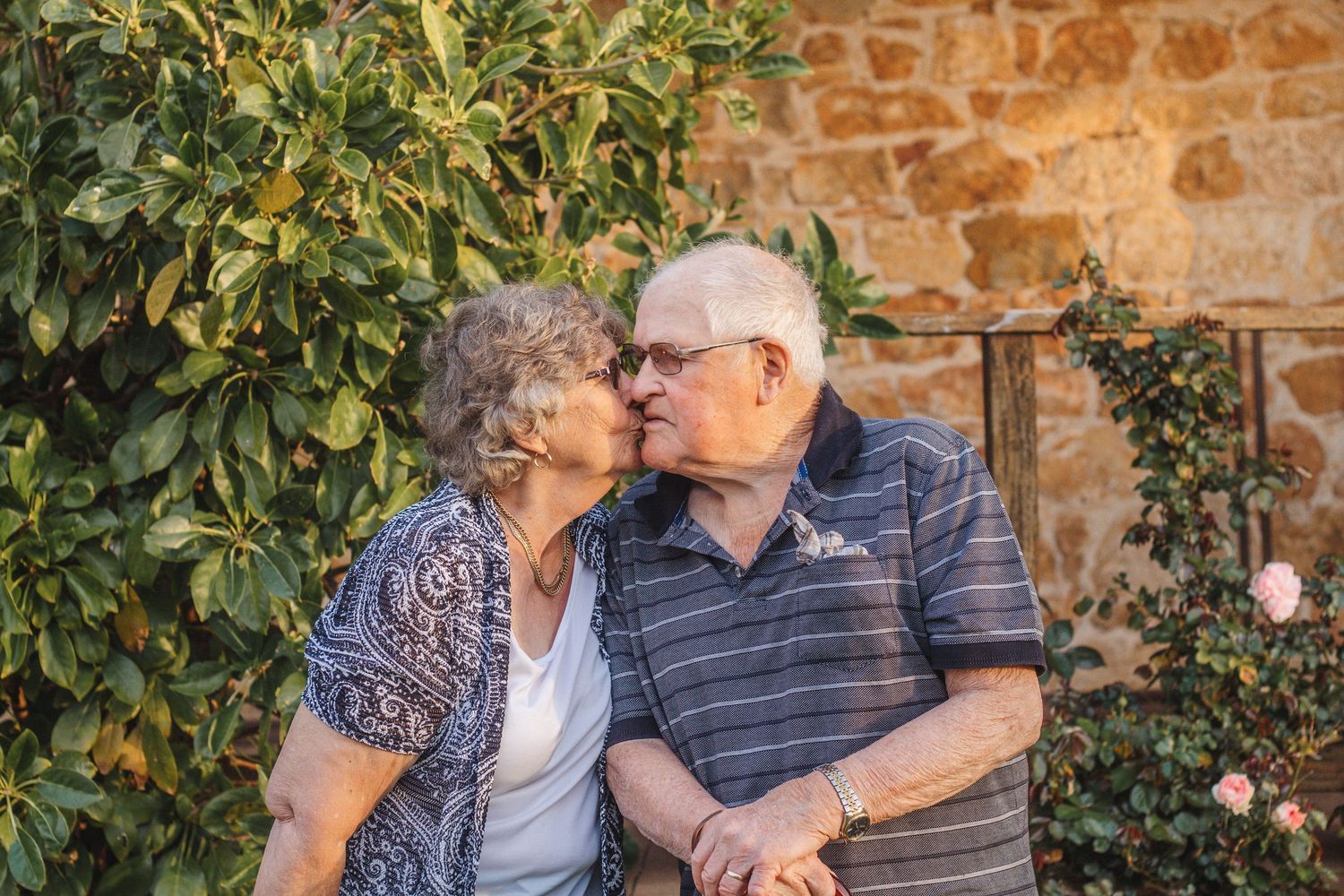 Loving elderly couple sharing an affectionate moment outdoors beside green foliage and stone wall.