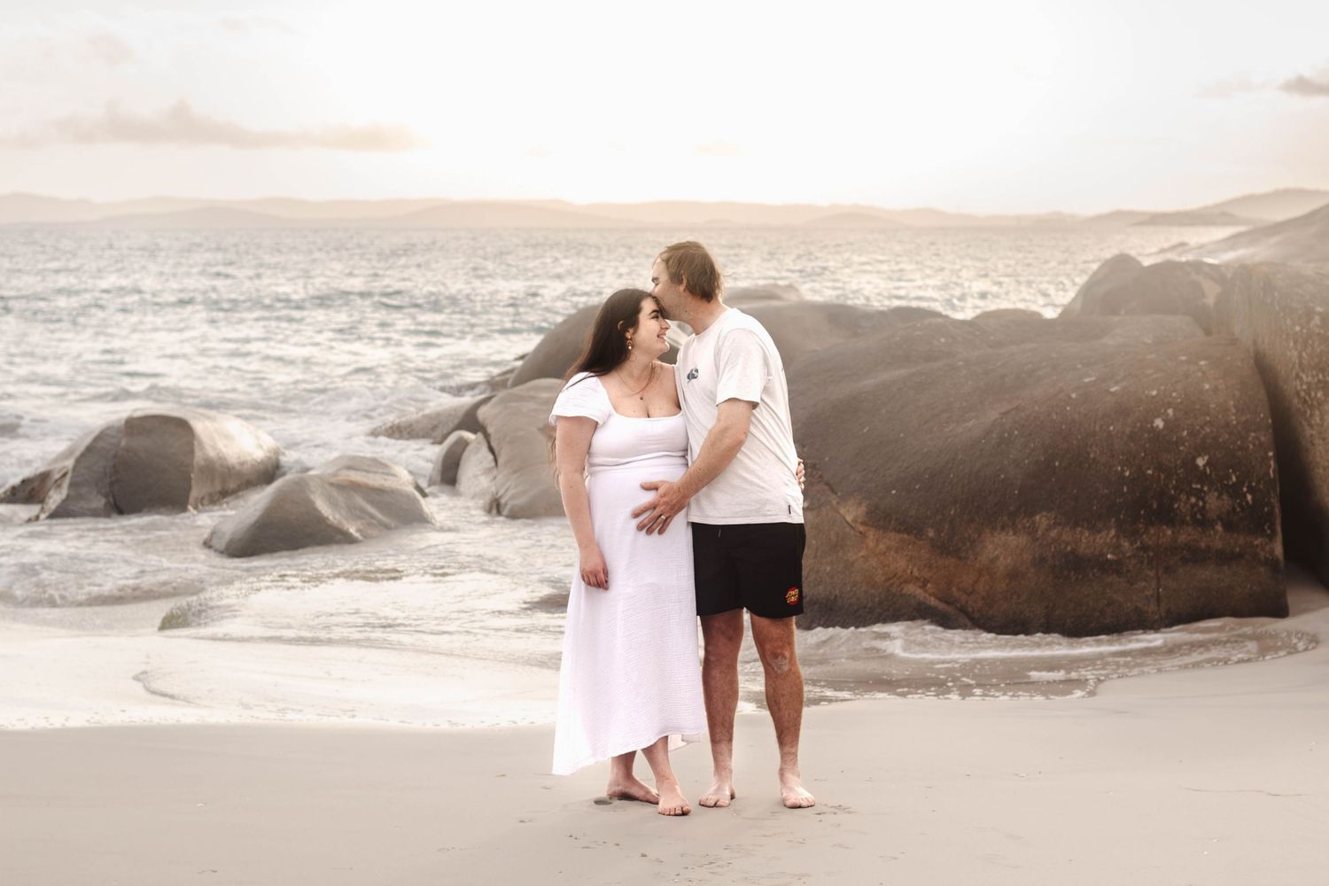 Couple in casual attire poses for maternity photo on beach with large rocks and ocean waves.
