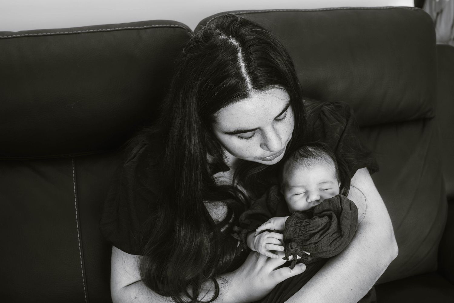 Mother in black shirt holding sleeping newborn close on leather couch in monochrome photo.